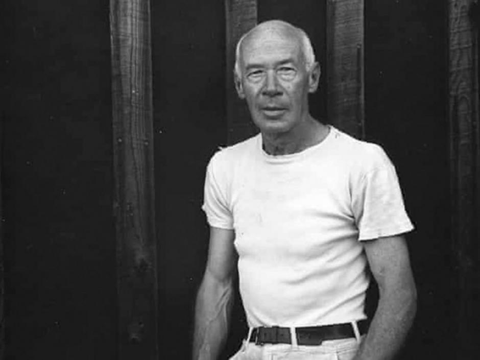 Black and white photo of Henry Miller, a slender and bald older white man, wearing a white tshirt and slacks.