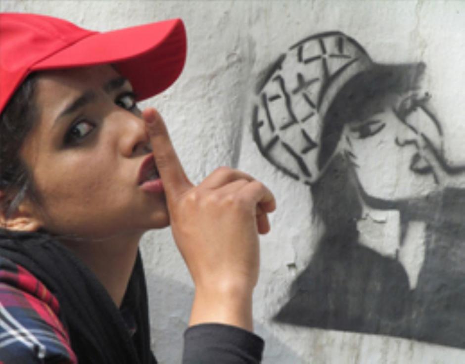 A young woman with a red baseball hat holds her finger to her lips. She's posing next to a street art portrait of herself in the same  clothes and pose.