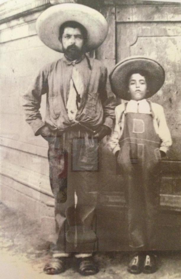 Man with dark hair and a beard and mustache, wearing informal clothes and a sombrero, standing next to a young boy in overalls and a sombrero.