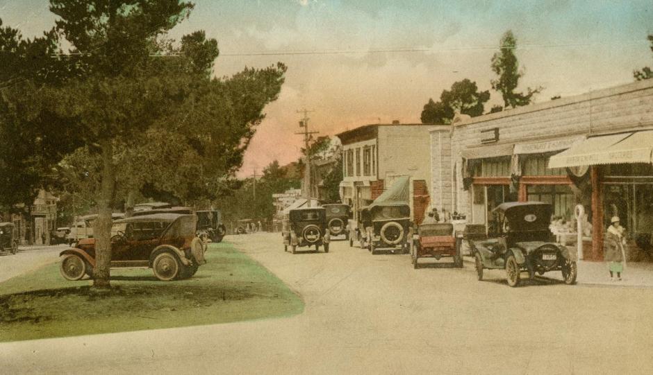 Old color-tinted postcard looking down Carmel's main street around 1915. There are storefronts long the right, and a grassy median with trees on the left. Old-fashioned cars are in the street and parked on the median,